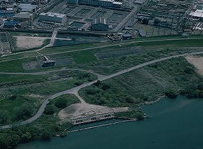 大きな川沿いに設備が作られ、道を挟んだ先に広い施設が建っている様子を上空から撮った写真