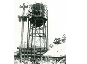 建物の隣に高く作られた配水塔が建っており、配水塔の下に数名の関係者が立っている白黒写真