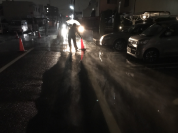 水が流れる道路にカラーコーンを置いている作業員の方が車のライトで照らされている写真