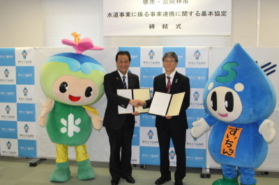 吉村市長と出耒管理者が協定書を持って握手し、その左右にトッピーとすいちゃんが立っている写真