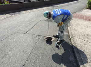 作業員の男性が道路上のマンホールの中に棒のようなものを差し込み調査を行っている様子の写真